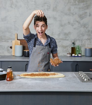 Eitan Bernath making pizza on a gray countertop and sprinkling cheese over the dough.