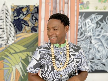 Kenzie Leon Perry, a Black man with short black hair, stands in front of verdant, tropical wallpaper display. He is seen from the chest up. He wears a black and white shirt that boasts swirling plant designs. The shirt has a lime green collar. He wears an oversized beige beaded necklace. His face is turned away from the camera, towards the left. He is grinning.
