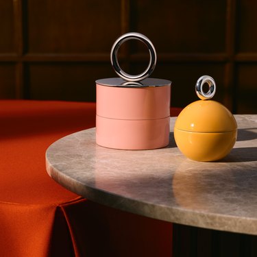 Two uncommon matters and H&M Home trinket boxes rest on a marble table. One is pink with a silver ring on the top, and the other is a yellow circular box, with a silver ring on top.