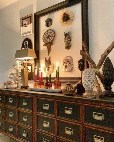 Perry's Kwanzaa display sits on a large chestnut and black console table. On the centerpiece is the tray with seven amber bottles, each holding white candles that are dipped in either red, green, or black. On the left side of the tray is a gaudy gold lamp that has a bust of a face affixed to the top, underneath the shade. Next to that is a small white Christmas tree, and in front of that are some decorative bowls. On the right side of the tray are collection of sculptures, some woven and others ceramic. These resemble eggs. Another sculpture is of an African mask. It is set up next to three pieces of driftwood. Behind the Kwanzaa display, on the wall, four frames. Three depict paintings. The largest frame holds six African masks. The masks depict a zebra, a sun face, and other fanciful characters.