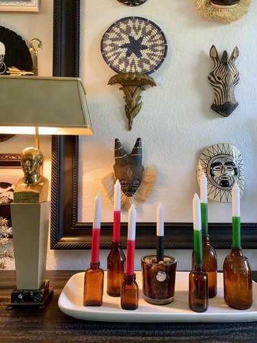 A close up of a Kwanzaa display. A gold lamp with a bust of a man attached at the top is arranged next to a tray. The tray has seven amber bottles, all varying sizes and widths. Each bottle has a candle placed inside. Three candles are white with red bottoms, one candle is white with a black bottom, and three remaining candles are white with green bottoms. An assortment of African masks hand in a frame hanging behind the table and display. The masks depict a zebra, a sun face, and other fanciful characters.