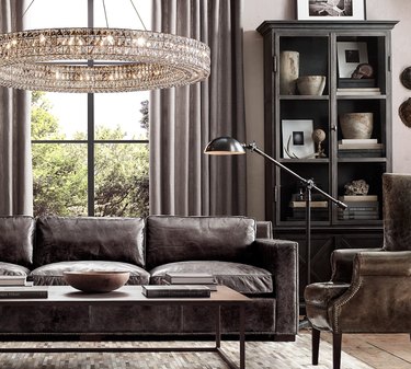 Industrial living room with dark leather couch and large round crystal chandelier, floor lamp, coffee table, leather wingback chair.