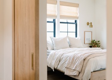 Bedroom with off white bedding, brass sconce, plant, windows, blinds.