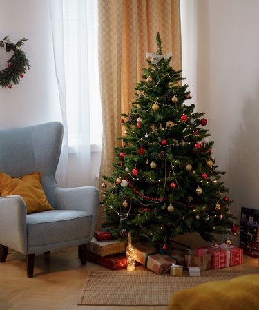christmas tree with lights and ornaments and presents underneath next to blue chair
