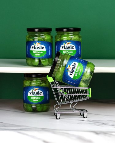 A jar of Vlasic pickles in a mini shopping cart next to another jar with two additional jars on a white shelf above in front of a green wall.