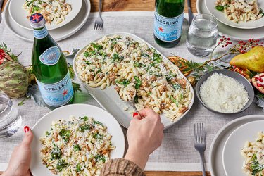 A holiday table is set with Gnocchetti con Salsiccia e Broccolini at the center of it. There is a white hand with red nail polish scooping pasta onto an individual plate. Two bottles of San Pellegrino rest on the table, with a. light grey table runner down the middle There is a centerpiece with red holly berries, a yellow pear, pomegranate, dried orange slides and an artichoke.