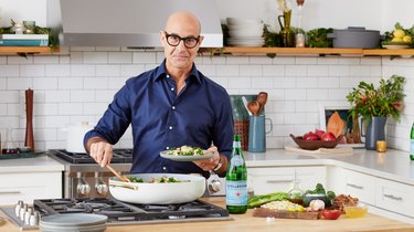 Actor Stanley Tucci wears a navy blue button down shirt and is standing in front of a stovetop with a plate of pasta in his hand. The white pot in front of him is filled with pasta, and his other hand holds a wooden spoon that's resting in the pot. There's a bottle of San Pellegrino on the counter next to him, as well as a wooden cutting board filled with ingredients for the pasta dish in the pot, including broccolini, olive oil, salt, garlic and pasta.