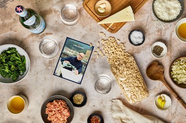 A flat lap of all of the ingredients for Gnocchetti con Salsiccia e Broccolini. The surface is a textured, light brown surface with a bowl of broccolini, a small dish of olive oil, a bottle of San Pellegrino, two glasses of the mineral water, a cutting board with cheese, a bag of dry pasta, small dishes with spices and cheese, a wooden spoon, garlic cloves, and a small picture of Stanley Tucci pouring a glass of San Pellegrino.