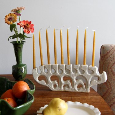 Ceramic menorah with yellow candles on a wooden table next to a vase of flowers.