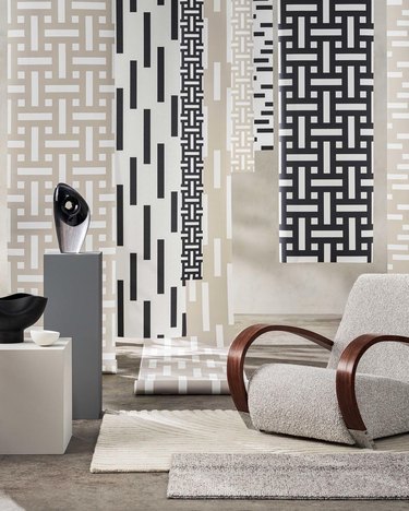 Patterned wallpaper borders hanging from the ceiling in a white room with a white chair and cream rug on a gray floor.