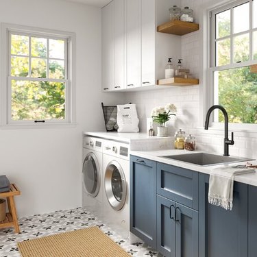 A laundry room with a side-by-side washer and dryer, open shelving, and a laundry room sink with blue cabinets