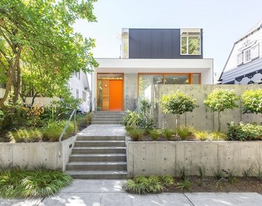 A contemporary house with an orange door and orange window panes. The square home sits on a small hill, with a long front pathway to the front door, surrounded by trees and small concrete walls.