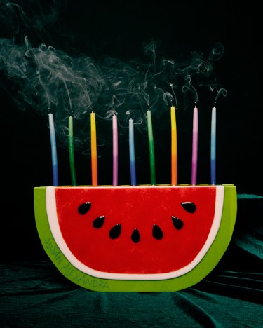 Watermelon-shaped menorah with colored candles on a black background.
