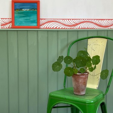 A green chair with a plant sitting in front of a green wall with a red and white wallpaper border.