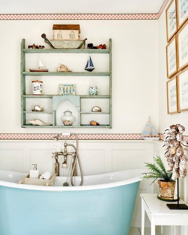 A bathroom with white walls, red wallpaper borders, a green wall shelf with trinkets, and a blue bathtub.