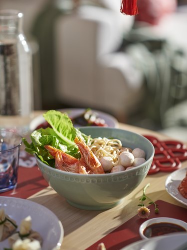 A light green bowl full of noodles, shrimp, and veggies on a wooden table surrounded by other dishware.