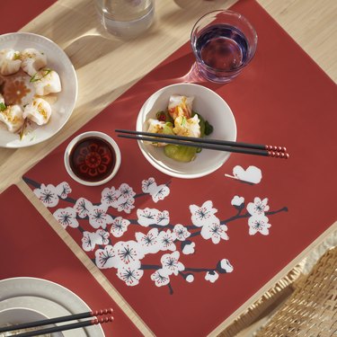A wooden table with a red cloth decorated with white flowers topped with a white bowl with black chopsticks.