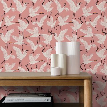 Pink wallpaper patterned with white cranes behind a light wood table topped with white sculptural vases.