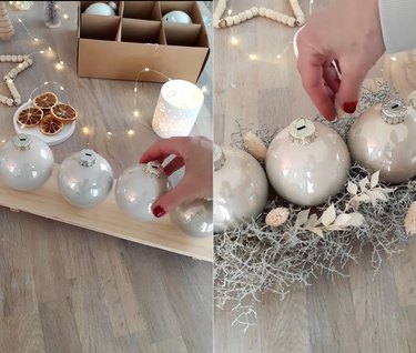 Split screen image of a hand putting a Christmas ornament on a plank of wood to the left and a hand placing foliage around the Christmas ornaments to the right