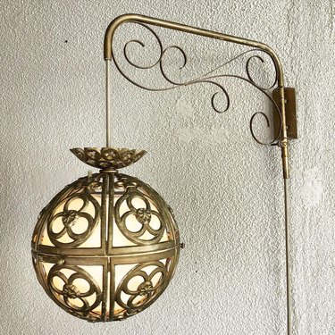 A gold Hollywood regency counter-weight lamp with filigree metalwork hanging off a bracket on a white wall.