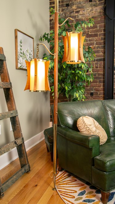 A midcentury teak tension pole lamp in the corner of an apartment in front of a red brick wall and next to a green leather couch.