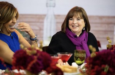 Ina Garten, wearing a black top and a purple scarf, sits next to Hoda Kotb at a holiday table on The Today Show