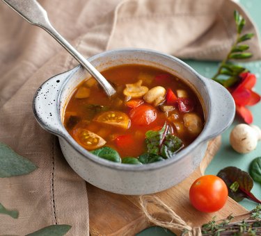 A bowl of vegetable soup with tomatoes and basil in a ceramic bowl with a silver spoon.