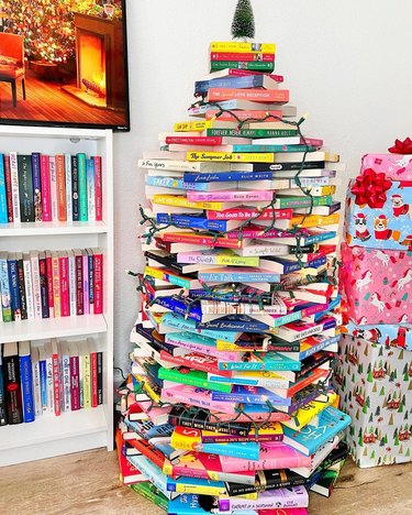 A rainbow Christmas book tree next to a stack of wrapped presents and a white bookshelf.