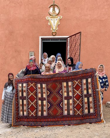 A group of Amazigh women holding up a red and white Moroccan rug in front of a terra cotta orange building.