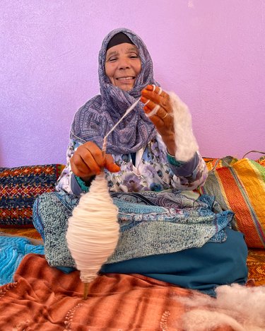 A woman sitting on a textile while turning wool into yarn in her hands.