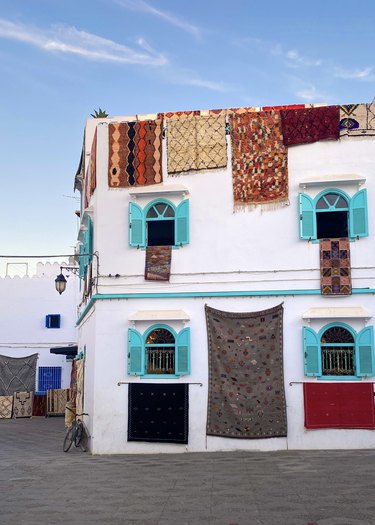 A white building with bright blue shutters that has Moroccan rugs hanging off the roof and out of the windows.