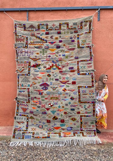 A woman standing behind a large Moroccan rug featuring colorful symbols in front of an orange building.