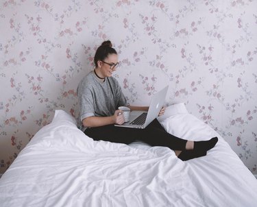 Woman in a gray top and black pants using a laptop on a white bed in front of a pink floral wall.