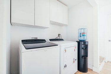 A top-loading washing machine and a dryer in a white laundry room next to a water cooler