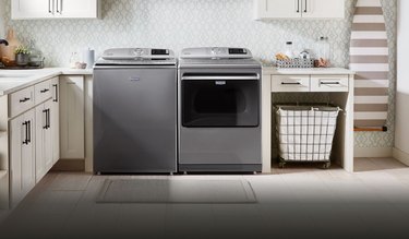A stainless steel washer and dryer combo from Maytag in a large laundry room with utility sink, ironing board, and a hamper