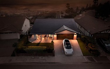 A ranch-style house at night with solar roof shingles; a Tesla is parked in front