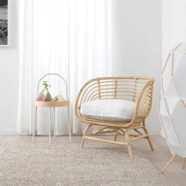 Rattan chair with a white cushion in a white room with a side table and oatmeal colored rug