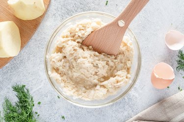 The grated potato, chopped onions, all-purpose flour, egg, salt, and pepper mixed with a wood spoon in a glass bowl.