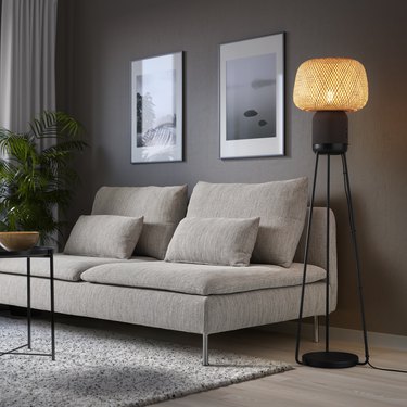 The IKEA Symfonisk floor lamp, which doubles as a speaker, in black with a bamboo shade, next to a beige couch with no arms.