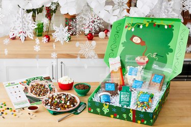 A festive HelloFresh Buddy the Elf Spaghetti meal kit is open on a kitchen counter, with paper snowflakes and red and green ornaments behind it. The recipe card is on the counter along with a bowl of spaghetti topped with candy and marshmallows, with a bowl of maple syrup, a bowl of marshmallows and a bowl of chocolate rice cereal to the side.
