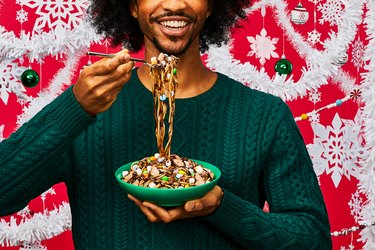 A black man in a green cable knit sweater smiles and holds a bowl of Buddy the Elf spaghetti in one hand, and a fork full of spaghetti in the other. The spaghetti is covered in chocolate syrup, candy and marshmallows, and the wall behind them is red, with white paper snowflakes, white tinsel, green ornaments and beaded garland.