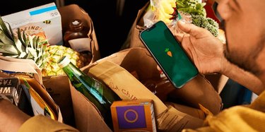 A black man holds a phone with the Instacart logo on the screen, amidst paper bags full of groceries.