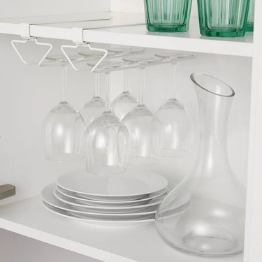 A white clip-on rack inside a white kitchen cabinet holding six clear wine glasses.