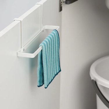 A white clip-on towel rack holding a blue striped tea towel on the inside of a lower white kitchen cabinet.