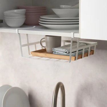 A white basket clipped onto the bottom of a white kitchen cabinet filled with plates and bowls.