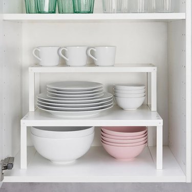 Two white shelf inserts stacked on top of each other in a kitchen cabinet, holding white and pink plates, bowls, and mugs.