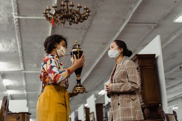 Two people in masks at an antique estate sale, with one person holding up a black and gold antique vase.