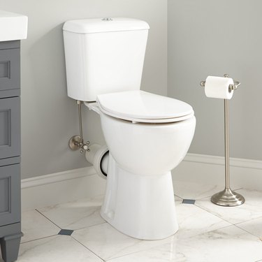A white dual flush toilet on a marble floor and a silver toilet paper holder
