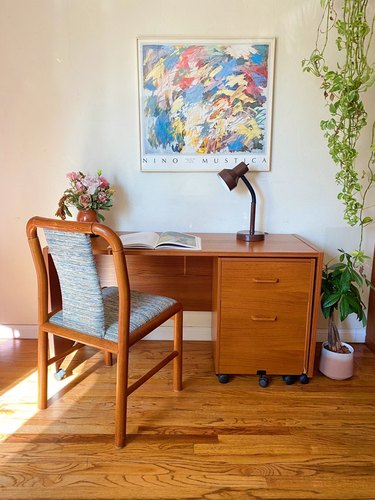 A Danish teak desk and matching upholstered chair topped with a vase of flowers and a brown vintage table lamp.
