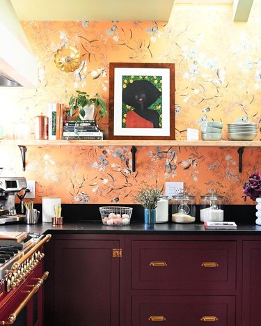Sunlit kitchen with yellow floral wallpaper, deep red cabinets, and framed portrait painting of an African-American woman against a green and yellow background.